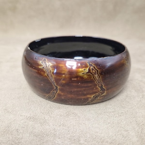 Puede incluir: Un cuenco redondo marrón con un acabado brillante y un interior negro. El cuenco tiene un patrón texturizado único con vetas doradas y marrones. El cuenco es un objeto decorativo.