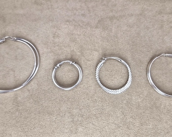 May include: A collection of four silver-toned hoop earrings arranged in a row. The set includes one large smooth hoop, one small smooth hoop, one small textured hoop with a braided pattern, and one medium-sized smooth hoop, all featuring hinged closures.