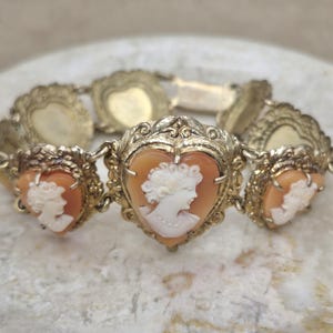 May include: A gold-toned metal bracelet featuring a series of heart-shaped links. Three of the links contain carved shell cameos depicting a profile of a person. The remaining links are solid gold-toned metal with a decorative scalloped border.