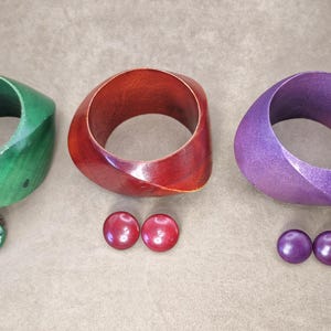 May include: Three wooden bangles in green, red, and purple, with matching stud earrings. The bangles have a geometric design. The earrings are round and shiny. The jewellery is displayed on a neutral surface.