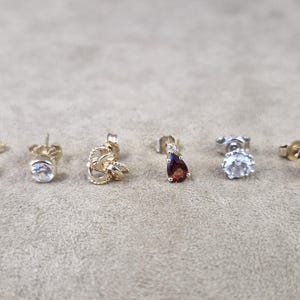May include: Six earrings in a variety of styles and designs. The earrings feature gold and silver metals, with gemstones in red, clear, and purple. Styles include studs and drop earrings, showcasing different shapes and settings.