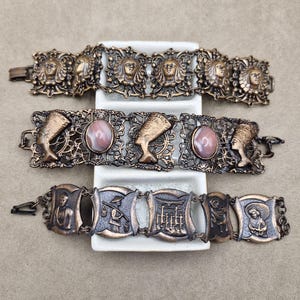 May include: Three antique bronze bracelets. The top bracelet features sculpted faces. The middle bracelet has Egyptian designs with oval pink stones. The bottom bracelet has religious and historical scenes.