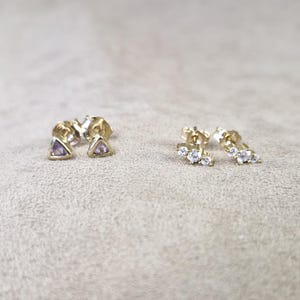 May include: Two pairs of gold earrings with small gemstones. One pair features a triangular purple stone, and the other pair has three small round clear stones.