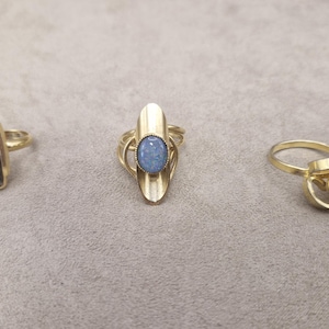 Puede incluir: Tres anillos de tono dorado. El anillo izquierdo presenta una cara rectangular con incrustaciones azul oscuro moteadas. El anillo central tiene un engaste dorado alargado y puntiagudo con una piedra azul ovalada. El derecho es geométrico.