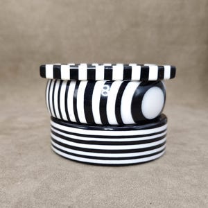 Könnte beinhalten: Ein Stapel aus vier schwarz-weiß gestreiften Armreifen. Der oberste Armreif hat ein Schachbrettmuster, während die anderen vertikale und horizontale Streifen aufweisen. Die Armreifen sind auf einem neutralen Hintergrund gestapelt.