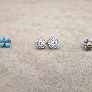 May include: Three pairs of earrings are displayed. The first pair features turquoise-coloured, four-petal flower designs. The second pair has clear, round stones surrounded by small, silver-coloured stones. The third pair is daisy-shaped with gold centres.
