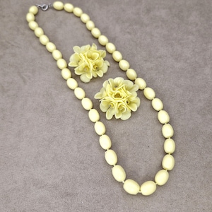 Könnte beinhalten: Eine cremefarbene Halskette mit ovalen Perlen und einem silbernen Verschluss, kombiniert mit zwei passenden floralen Broschen. Das Schmuckset wird auf einem neutralen Hintergrund präsentiert, der das elegante Design und die Farbe hervorhebt.