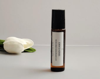 May include: A small, brown glass roll-on bottle with a black cap. The label reads "Erin's Herbs Rose and Sandalwood". A white flower and green leaves are in the background. The bottle contains 10ml of product.