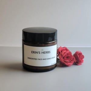 May include: A brown glass jar with a black lid, labeled "ERIN'S HERBS UNSCENTED FACE AND HAND CREAM" with a green star logo. The jar is 4 fl oz / 118 ml. Red roses are next to the jar.