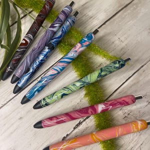 May include: A collection of colorful, marbled-design pens. The pens feature a variety of colors, including red, purple, blue, green, pink, and orange, with silver accents. The pens are arranged on a light-colored wooden surface with green decorative accents.