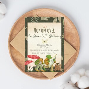May include: A birthday invitation with the text "HOP ON OVER for Bonnie's 5th Birthday". The invitation features illustrations of frogs, mushrooms, and foliage. The invitation is on a wooden plate with a brown envelope.