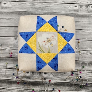 May include: Square pincushion with a patchwork star design in blue and yellow, centered with a dragonfly illustration. The pincushion is beige with a floral pattern. Several pins with colorful heads are inserted into the cushion.