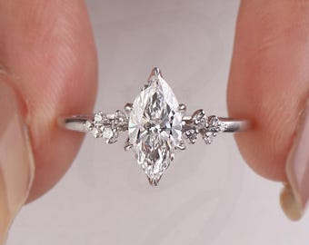 Op de afbeelding: Een verlovingsring met een marquise-geslepen diamant en kleinere accentstenen. De ring heeft een zilveren band en wordt tussen twee vingers gehouden. Het logo van The Bride Jewelry is rechtsonder zichtbaar.
