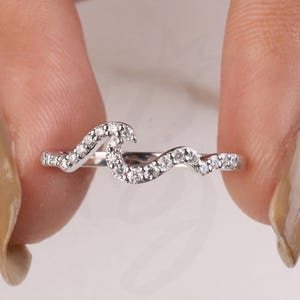 May include: A delicate, wave-shaped ring with a silver band, adorned with small, sparkling clear stones. The ring's design is elegant and minimalist, perfect for everyday wear or special occasions. The Bride Jewelry logo is visible in the bottom right corner.