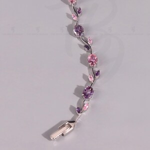Puede incluir: Una pulsera de tono plateado con un diseño de enredadera que presenta gemas ovaladas alternas en rosa y morado. La estructura metálica incluye pequeños detalles en forma de hoja y un cierre rectangular en un extremo. Logo de The Bride Jewelry.