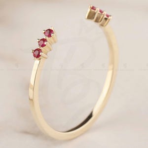 May include: A gold-colored open ring with three small, round, red gemstones on each end. The ring is set against a soft, neutral background, highlighting the jewelry's delicate design and vibrant gemstones.