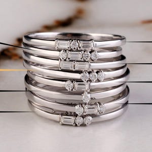 Puede incluir: Pila de anillos de plata con pequeñas piedras preciosas transparentes. Algunos anillos tienen una fila de piedras de talla baguette, mientras que otros presentan piedras redondas. Los anillos se muestran sobre una superficie de mármol blanco y gris. Logotipo de The Bride Jewelry.