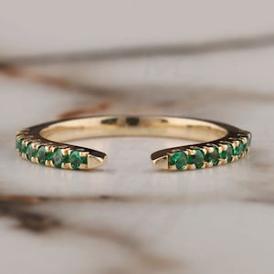 May include: A gold ring with a row of emerald green gemstones set in a delicate band. The ring is open on one side, making it a half eternity band.