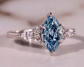 May include: A marquise-cut, light blue gemstone ring with a silver band. The ring features smaller, round, clear gemstones on either side of the main stone. The Bride Jewelry logo is visible in the bottom right corner.