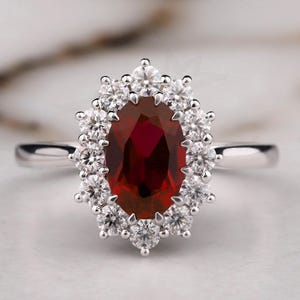 May include: A silver ring featuring a large, oval-cut, deep red gemstone surrounded by a halo of smaller, clear gemstones. The ring's band is thin and polished, and the setting is intricate, creating a sparkling effect. The Bride Jewelry logo is visible.