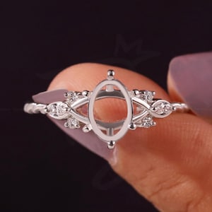May include: A silver-toned ring setting featuring a central oval-shaped prong mount for a gemstone. The band is adorned with small, clear, round-cut stones set in a delicate, symmetrical metal framework that extends from the central setting.