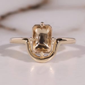 May include: A gold-colored ring with an emerald-cut setting. The ring features a unique design with a curved band and decorative elements around the setting. The Bride Jewelry logo is visible in the bottom right corner.