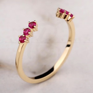 Puede incluir: Un anillo abierto de color dorado con tres vibrantes piedras preciosas rojas en un lado y un grupo de piedras preciosas rojas en el otro. El anillo tiene un diseño delicado y curvado, mostrando las piedras preciosas.