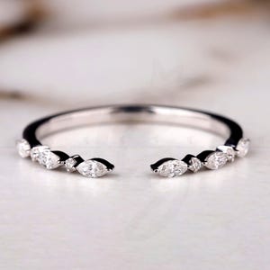 May include: A silver-toned open ring with a row of marquise-cut gemstones. The ring has a gap in the center and is set against a soft, blurred background. The Bride Jewelry logo is in the bottom right corner.