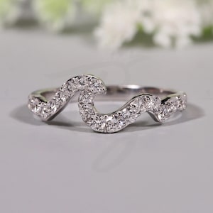 May include: A silver-toned ring with a wave-like design, encrusted with small, sparkling clear stones. The ring's curved shape is accentuated by the placement of the stones, creating a continuous, flowing pattern. The Bride Jewelry logo is visible.
