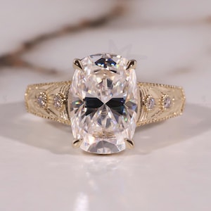 May include: A gold engagement ring with a large, oval-cut diamond as the centerpiece. The band features intricate filigree detailing and small accent diamonds. The ring is set against a neutral background.