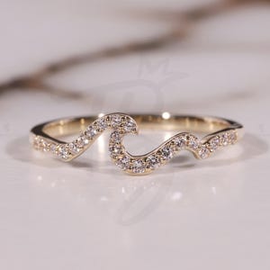 Può includere: Un anello color oro con un design a onda, incastonato con piccole gemme trasparenti. L'anello è posto su una superficie di marmo bianco. Il testo "THE BRIDE JEWELRY" è visibile in basso a destra.
