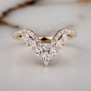 May include: A gold-toned ring with a V-shaped design. The ring is adorned with multiple clear, sparkling gemstones, including marquise-cut and round-cut stones. The ring is set against a soft, neutral background.
