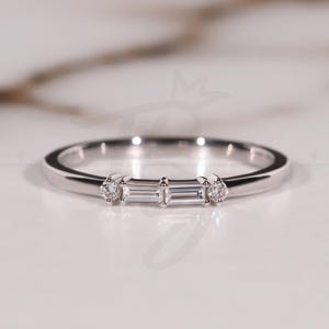 Könnte beinhalten: Ein zarter Silberring mit drei Edelsteinen. Der Mittelstein ist ein rechteckiger Baguetteschliff, flankiert von zwei runden Diamanten. Der Ring liegt auf einer weißen Marmoroberfläche.