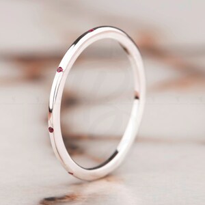 May include: A thin silver band with four small red gemstones set into the metal. The ring is on a light brown surface.