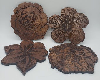 May include: Four wooden coasters with floral designs. The coasters are cut from dark brown wood and feature detailed engravings of a rose, hibiscus, and other flowers. The coasters are designed to protect surfaces from drink rings.
