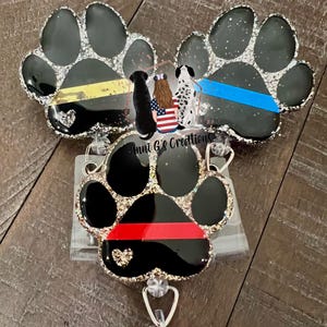 May include: Three paw-shaped badge reels with glitter edges. One has a yellow stripe, another a blue stripe, and the third a red stripe. Each paw has a heart detail. The text "Anni G's Creations" is visible.