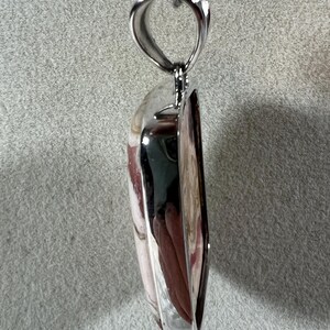 May include: A pendant necklace featuring a polished, heart-shaped stone with a silver-colored metal frame. The stone has a mix of pink, white, and brown hues. The necklace hangs from a delicate silver chain, creating an elegant accessory.