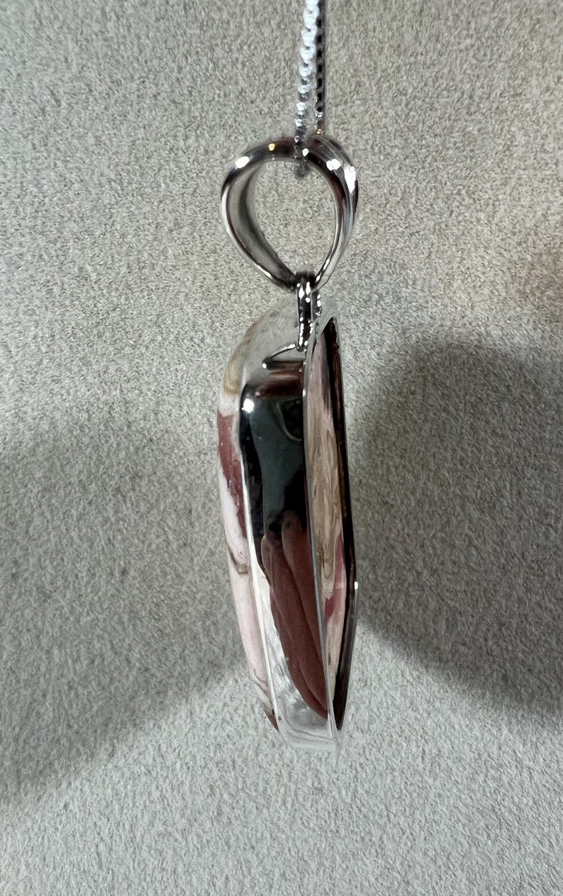 May include: A pendant necklace featuring a polished, heart-shaped stone with a silver-colored metal frame. The stone has a mix of pink, white, and brown hues. The necklace hangs from a delicate silver chain, creating an elegant accessory.