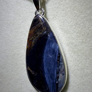 May include: A teardrop-shaped pendant with a polished, dark blue gemstone, accented with streaks of brown and gold. The stone is set in a silver-colored bezel and hangs from a delicate silver chain. The pendant is approximately 2 inches long.