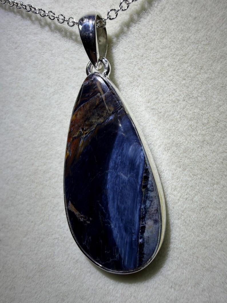 May include: A teardrop-shaped pendant with a polished, dark blue gemstone, accented with streaks of brown and gold. The stone is set in a silver-colored bezel and hangs from a delicate silver chain. The pendant is approximately 2 inches long.