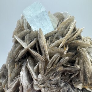 May include: A large, light blue aquamarine crystal sits atop a cluster of gray, mica-like crystals.