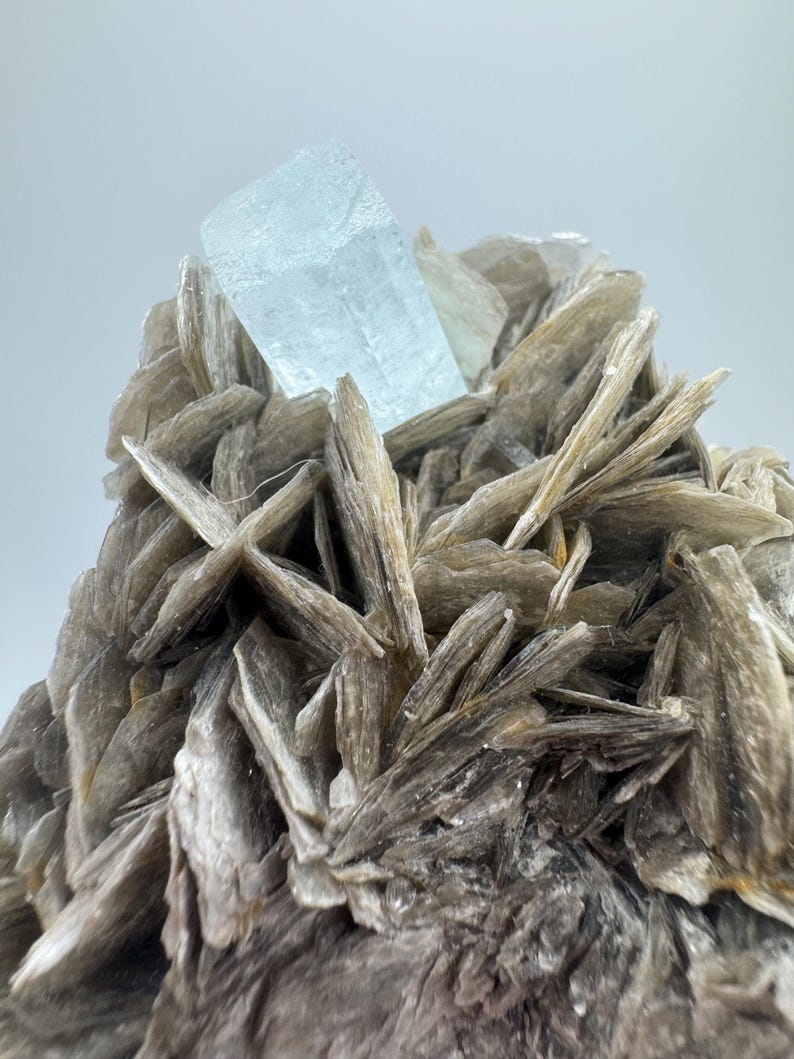 May include: A large, light blue aquamarine crystal sits atop a cluster of gray, mica-like crystals.