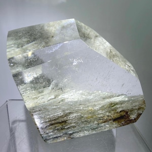 May include: A large, clear quartz crystal with a layered, textured base. The crystal has a geometric shape with sharp angles and a polished, transparent appearance. The base features earthy tones of brown and green.