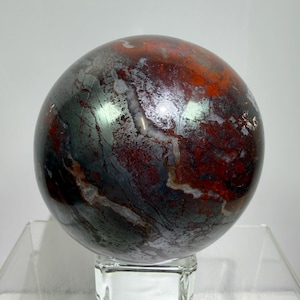 May include: A polished spherical stone featuring a marbled pattern of deep red, metallic grey, and white veins. The sphere is displayed resting on a clear, square glass stand, showcasing a smooth, reflective surface with varied mineral textures.