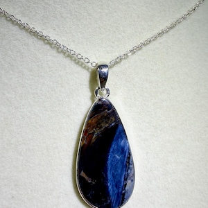 May include: A teardrop-shaped pendant with a silver-colored bezel and chain. The pendant features a polished gemstone with a mix of dark blue, brown, and white colors. The chain is delicate and hangs from the top of the pendant.