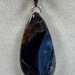 May include: A teardrop-shaped pendant with a polished, dark blue gemstone. The stone has streaks of brown and gold, set in a silver-colored metal frame. The pendant hangs from a delicate silver chain, creating a striking piece of jewelry.