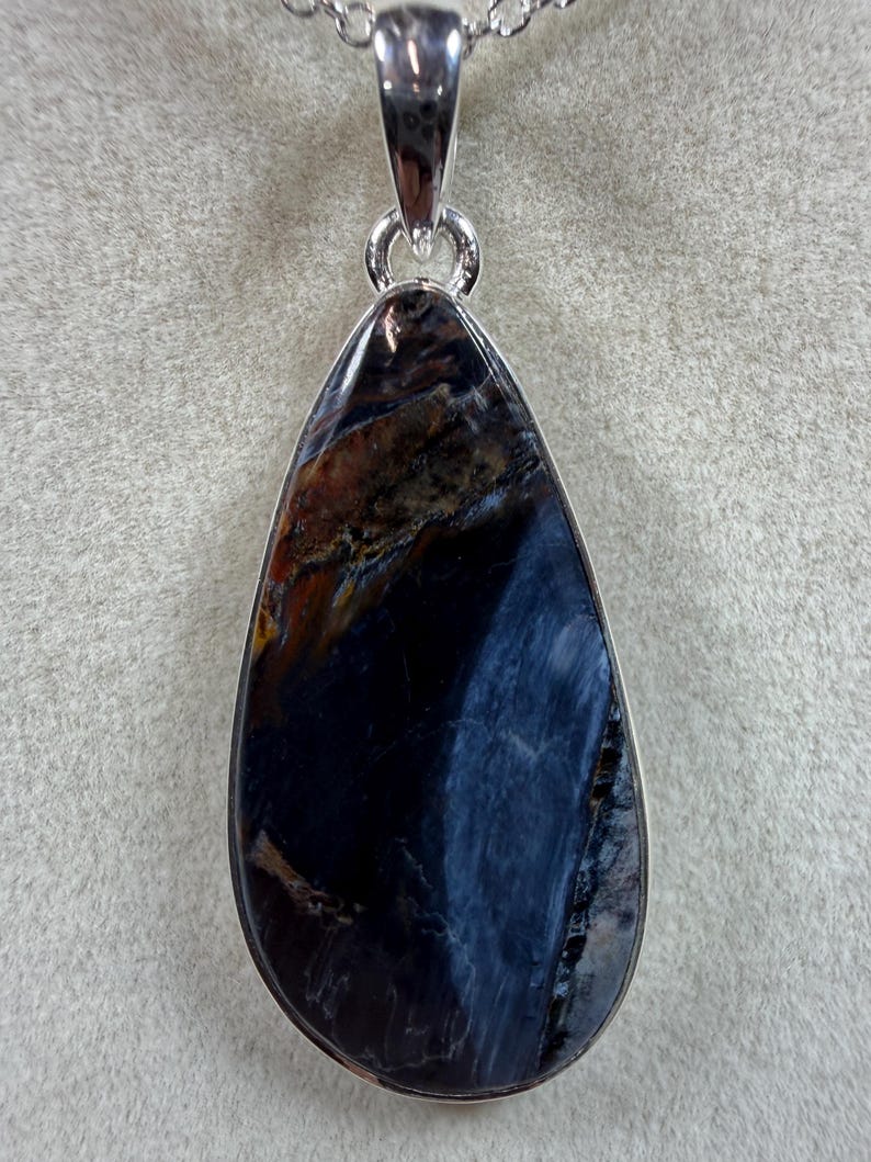 May include: A teardrop-shaped pendant with a polished, dark blue gemstone. The stone has streaks of brown and gold, set in a silver-colored metal frame. The pendant hangs from a delicate silver chain, creating a striking piece of jewelry.