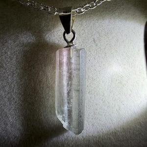 May include: A clear, elongated, raw crystal pendant suspended from a silver-toned metal chain. The crystal features a vertical, prismatic structure with subtle pinkish hues near the top bail, showcasing natural facets and a translucent appearance.