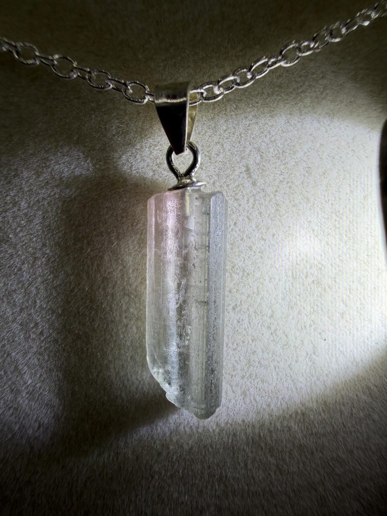 May include: A clear, elongated, raw crystal pendant suspended from a silver-toned metal chain. The crystal features a vertical, prismatic structure with subtle pinkish hues near the top bail, showcasing natural facets and a translucent appearance.