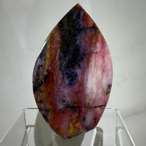 May include: A teardrop-shaped gemstone with a mix of colors, including pink, red, yellow, and black. The stone has a polished surface and is displayed on a clear acrylic stand. The gemstone's pattern is unique.